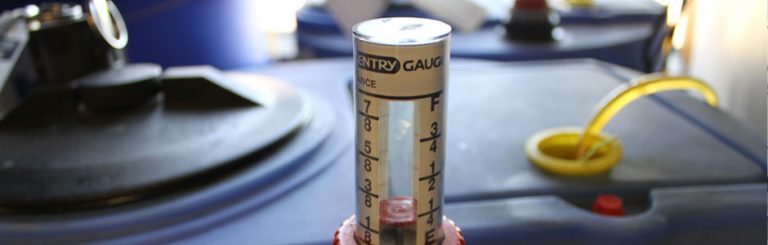 Close-up photo of a tank gauge with fractional markings and "ENTRY GAUGE" visible, next to a yellow hose.