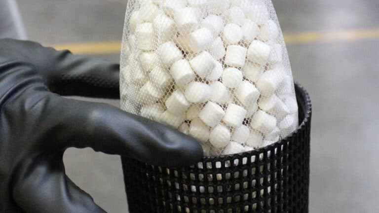 A gloved hand holding a mesh bag filled with small white controlled-release chemical tablets inside a black basket.