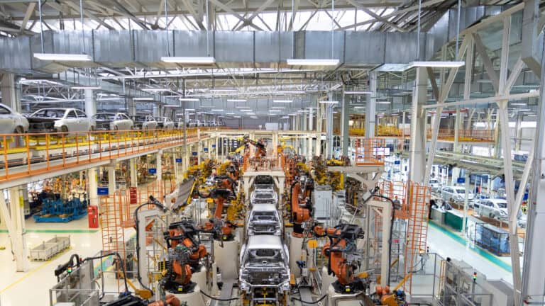 An automated car manufacturing factory floor with robotic arms assembling vehicle chassis along an industrial assembly line.