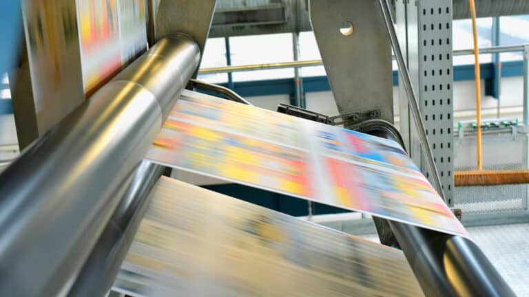 A high-speed industrial printing press running colorful printed paper through metal rollers in a printing facility.