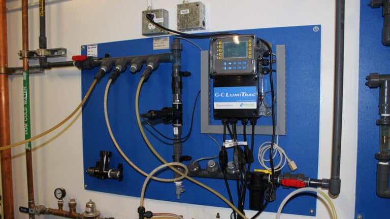 Wall-mounted G-C LumiTrac controller unit with digital display and connected piping on a blue panel in a mechanical room.
