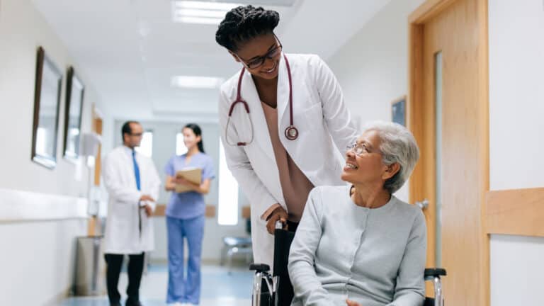 An elderly woman in a wheelchair smiles up at a doctor in a hospital hallway, with staff in the background.