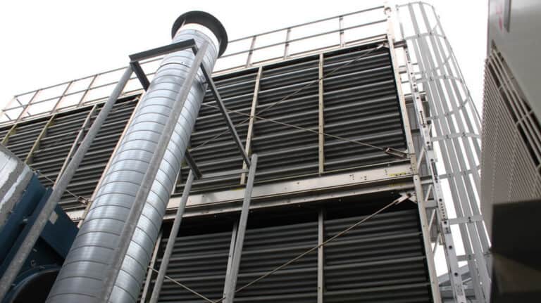 Low angle view of a large industrial cooling tower with corrugated metal louvers, a tall exhaust pipe, and structural framework.
