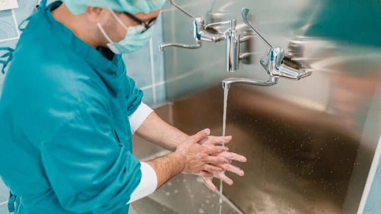 Healthcare worker in teal scrubs and a face mask washing soapy hands under running water at a stainless steel sink.