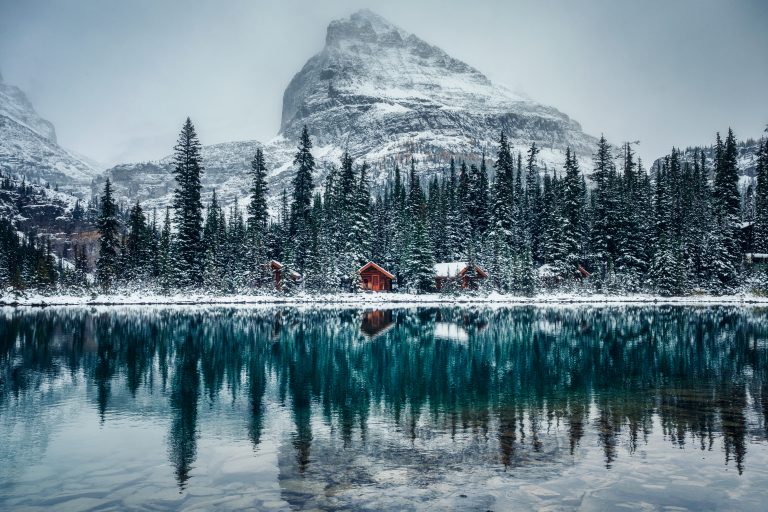 Snowy mountain lake reflecting pine trees and cabins, with a majestic snow-capped peak in the background.