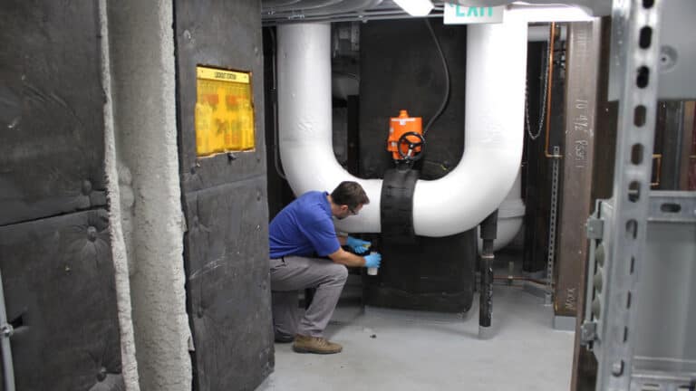 Technician in a blue shirt taking a water sample from large insulated pipes inside an industrial mechanical room.