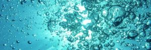 A close-up, underwater view of numerous bubbles rising in clear blue water towards a bright light source at the surface.