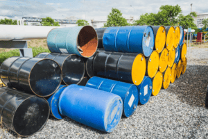 A stack of colorful barrels, primarily blue, yellow, and black, is arranged on gravel in the outdoors