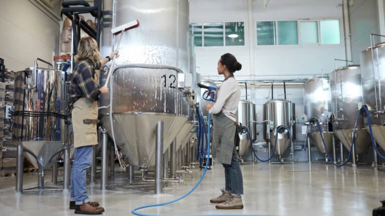 A man and woman in aprons wash a soapy stainless steel brewery tank with a long brush and a hose.