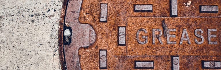 A close-up of a rusty, circular metal grease trap cover embedded in concrete, with the word "GREASE" clearly visible.