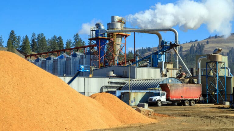 Industrial lumber mill facility with large piles of sawdust, processing machinery emitting steam, and a semi-truck parked under a clear sky.