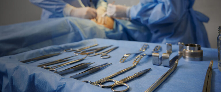 Carefully arranged clean metal surgical instruments inside a metal tray with surgery in the background.