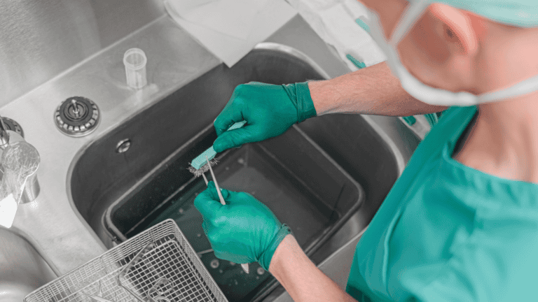 Person in green scrubs & gloves cleaning medical instruments in a sink with a brush, focusing on hygiene.