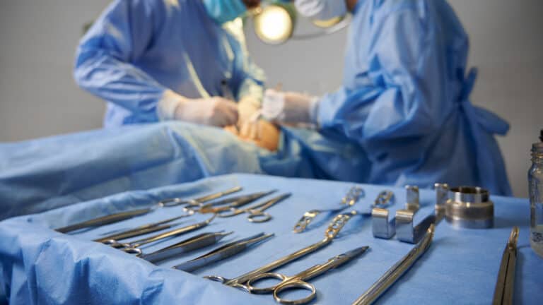 Carefully arranged clean metal surgical instruments inside a metal tray with surgery in the background.