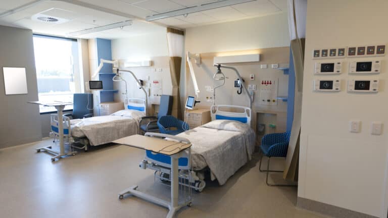 A modern hospital room with two adjustable patient beds, medical equipment arms, overbed tables, and wall-mounted controls.