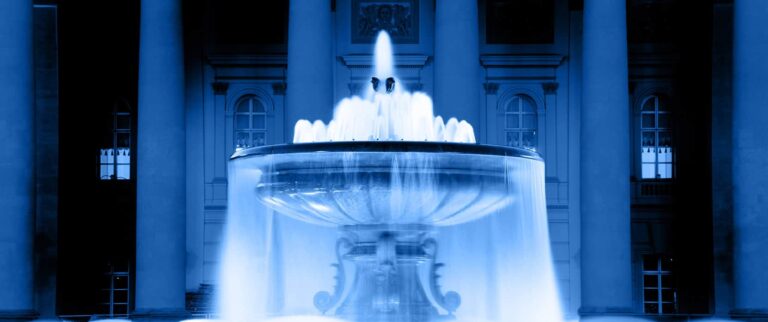 A scene with a decorative water fountain outside a building at night stylized with a blue and sepia color filter.