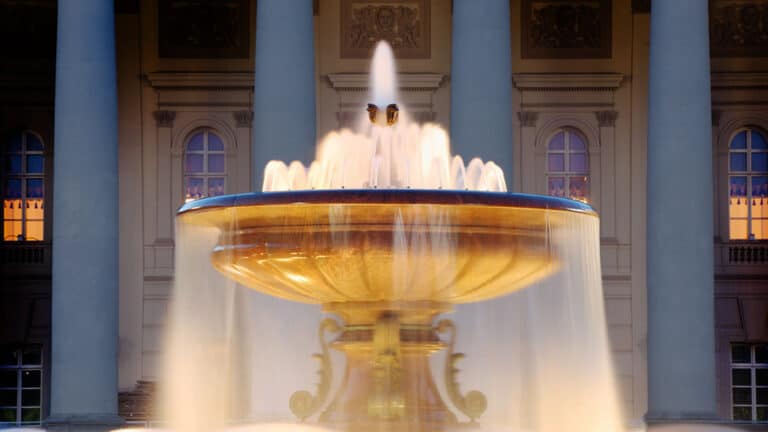 An illuminated ornate stone fountain spraying water in front of a classical building with large columns.