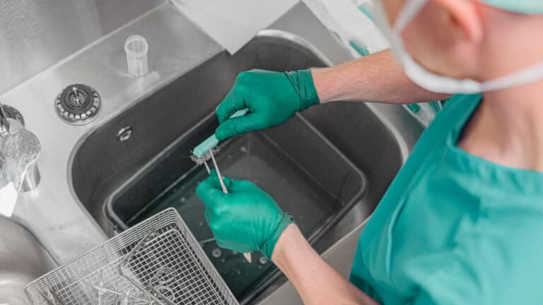 Medical worker in green gloves cleaning surgical instruments with a small brush over a stainless steel hospital sink.